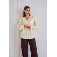 Baumwoll-Kaschmir-Crinkle-Bluse Frau steht in heller Baumwoll-Kaschmir-Bluse und braunen Hosen, an einer Wand.