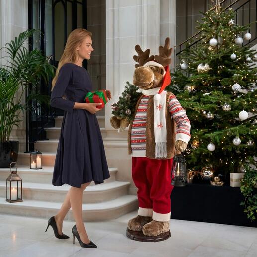 Tanzender Elch in festlicher Kleidung, hält ein Geschenk und steht vor einem Weihnachtsbaum.