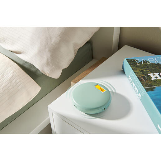 Vibrationswecker in sanftem Grün, auf einem Nachttisch neben einem Buch und einer Bettdecke.