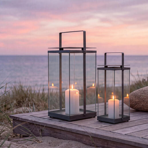 Zwei Design-Laternen auf einer Holzterrasse am Strand bei Sonnenuntergang.
