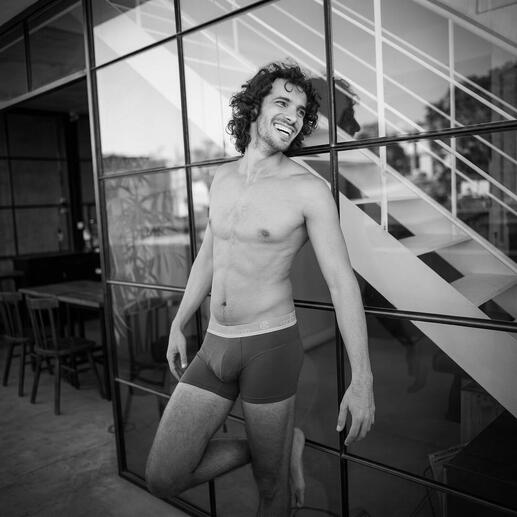 Mann in Boxershorts, lächelt vor Glaswand.