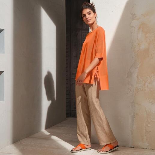 Frau in orangefarbenem Oberteil und Zehensandalen, posed in minimalistischem Raum