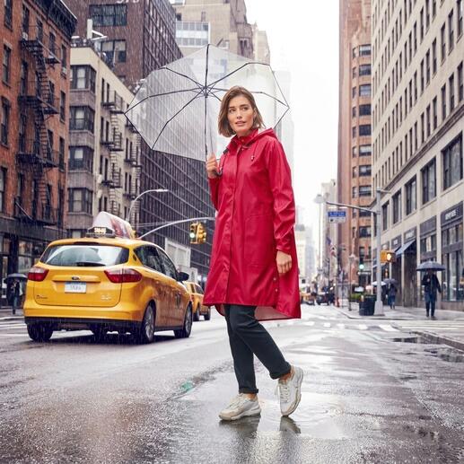 Frau in rotem Regenmantel hält einen durchsichtigen Regenschirm in einer urbanen Umgebung.