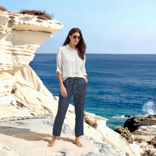 Frau in luftiger Bluse und Hose steht am Meer.