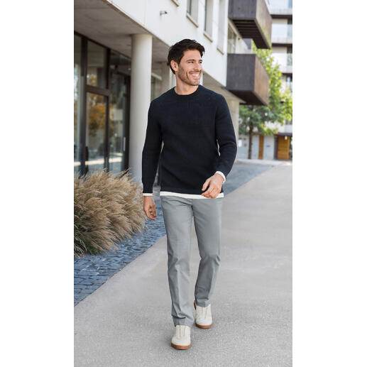 Hiltl Oxford-Chino Mann in einem schwarzen Waffel-Pullover und grauen Hosen, geht auf einem modernen Gehweg.