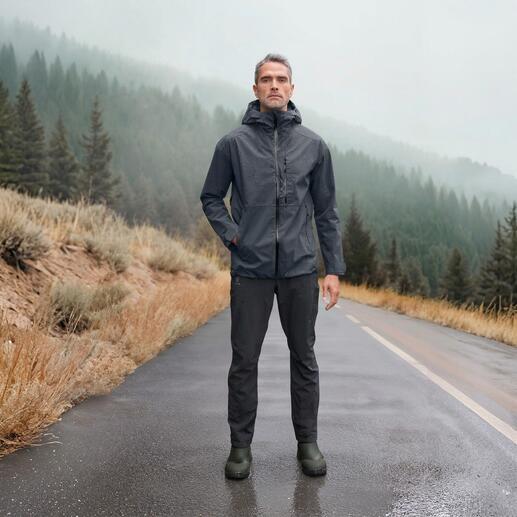 Mann in grauer Hardshell-Jacke steht auf nasser Straße, im Hintergrund Berge.