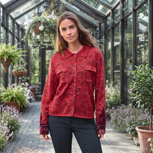 Frau in roter Jacquard-Strickbluse mit floralem Muster in einem Gewächshaus.
