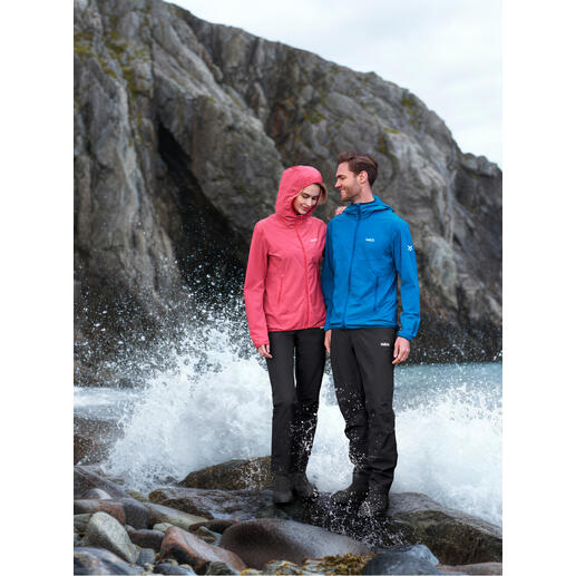Paar in Outdoor-Bekleidung, eine Person trägt pinke Softshelljacke, andere in blauer Jacke neben stürmischem Wasser.