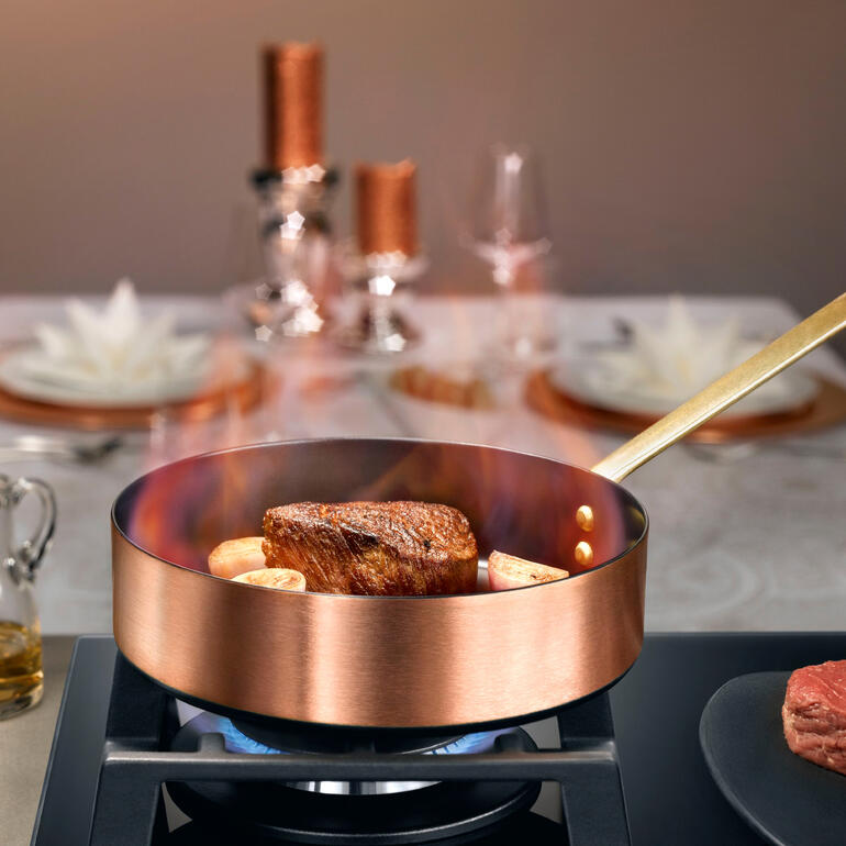 Induktions-Kupfer-Kochgeschirr auf dem Herd mit Fleisch und Gemüse.