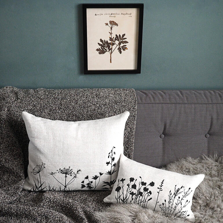 Zwei Leinenkissen mit schwarzen Pflanzenmustern auf einer Couch, ein Bild hängt darüber.