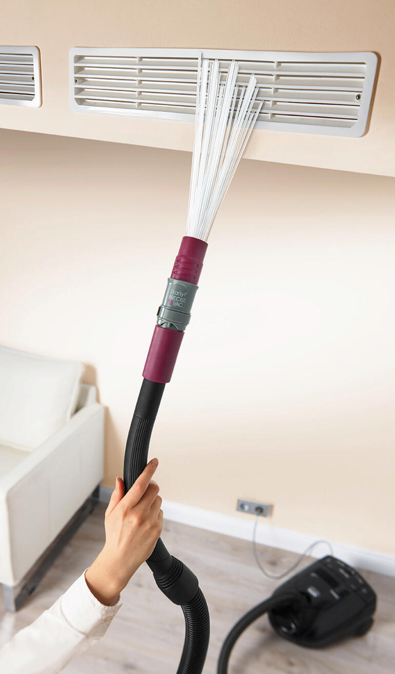 Hand mit Staubsaugeraufsatz, der eine Lüftungsschlitze reinigt.