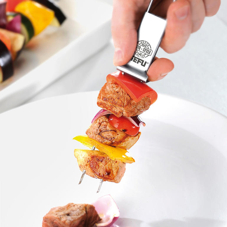 Hand mit einem Doppel-Grillspieß, der mit Gemüse und Fleisch belegt ist
