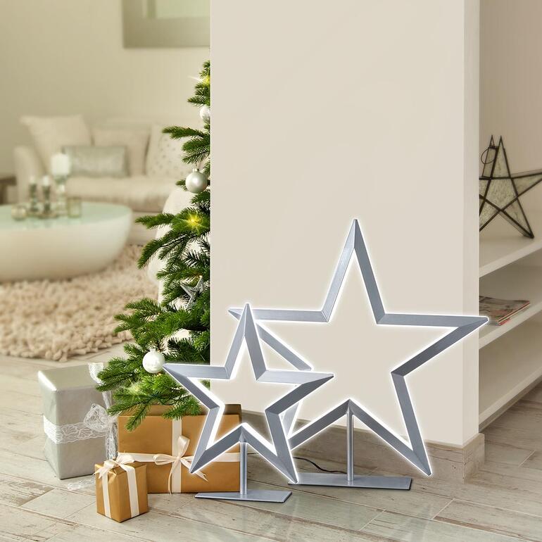 Zwei LED-Sterne neben einem Weihnachtsbaum in einem hellen Raum.