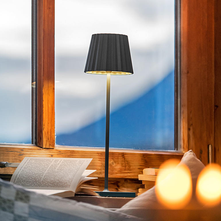 Akku-Tischleuchte in Schwarz, auf einem Tisch neben einer Fensterbank.