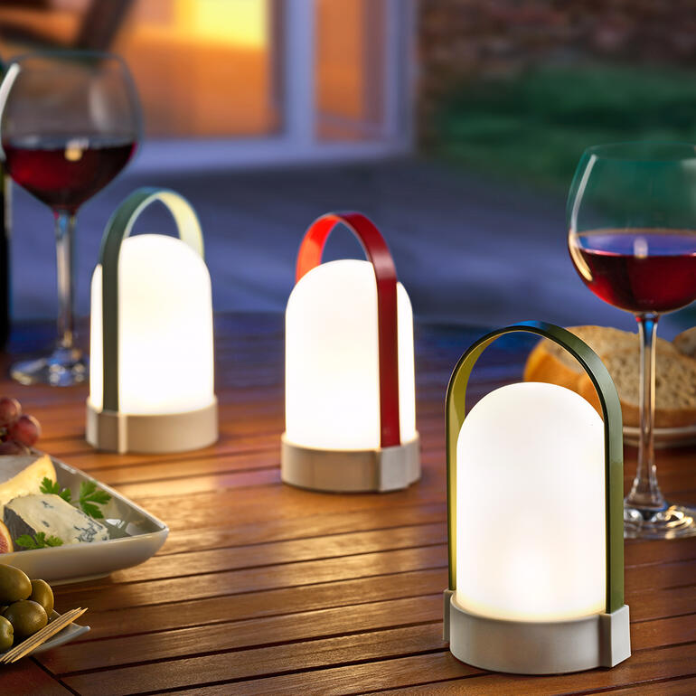 Drei Akkuleuchten Piccolos auf einem Tisch im Freien, umgeben von Speisen.