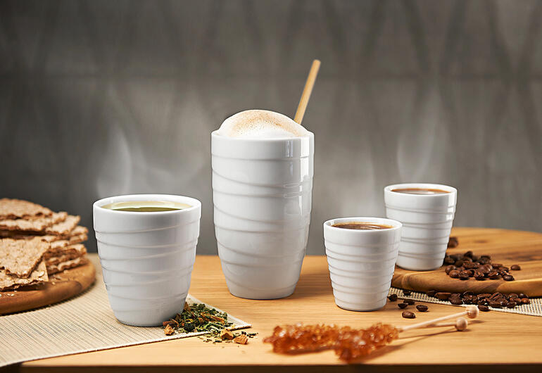 Vier Porzellan-Thermobecher auf einem Tisch, umgeben von Snacks.