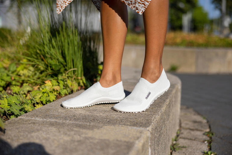 Weißer Barfuß-leguano® Slipper auf dem Boden, mit einer Frau im Hintergrund.