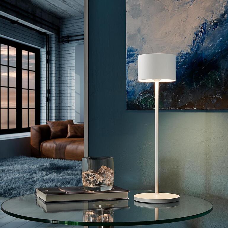 Tischleuchte in Weiß auf einem Tisch mit Kunstwerk im Hintergrund.