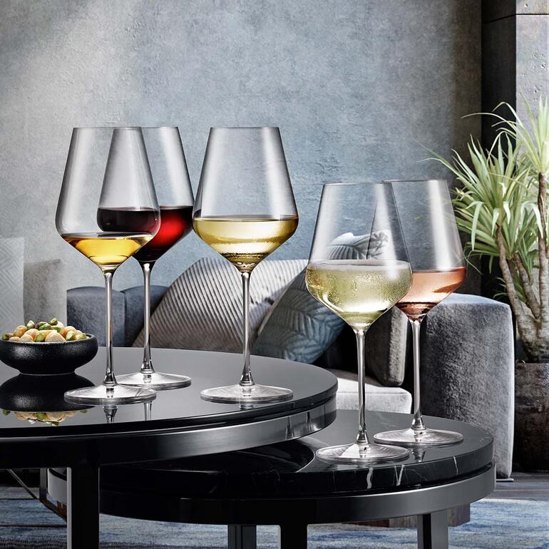 Vier Weingläser verschiedener Größen auf einem Tisch mit Snacks im Hintergrund.