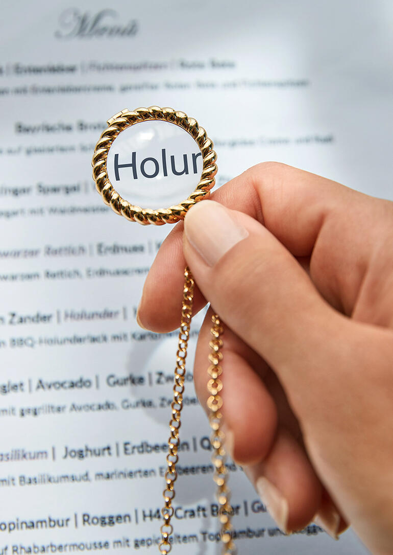 Hand hält Lupe an einer goldenen Kette über einem Menü.