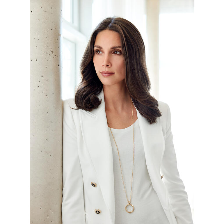 Die elegante Kette mit glasklarem Schmuckstein ist zugleich eine 6-fach vergrößernde Lupe. Frau in weißem Blazer trägt eine Lupenkette um den Hals.