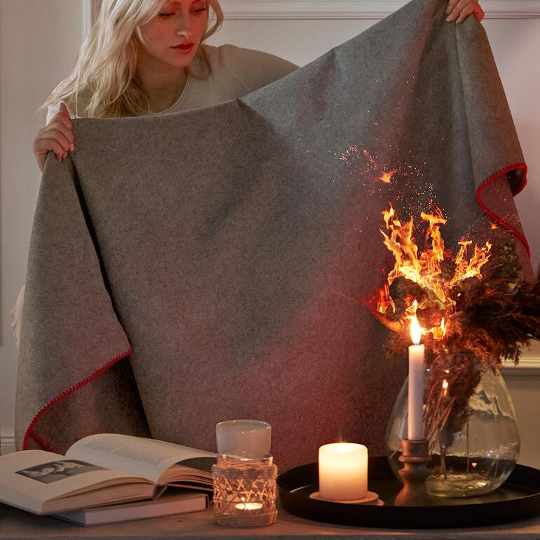 Frau hält eine Feuerlöschdecke vor sich, während Flammen im Hintergrund sichtbar sind.
