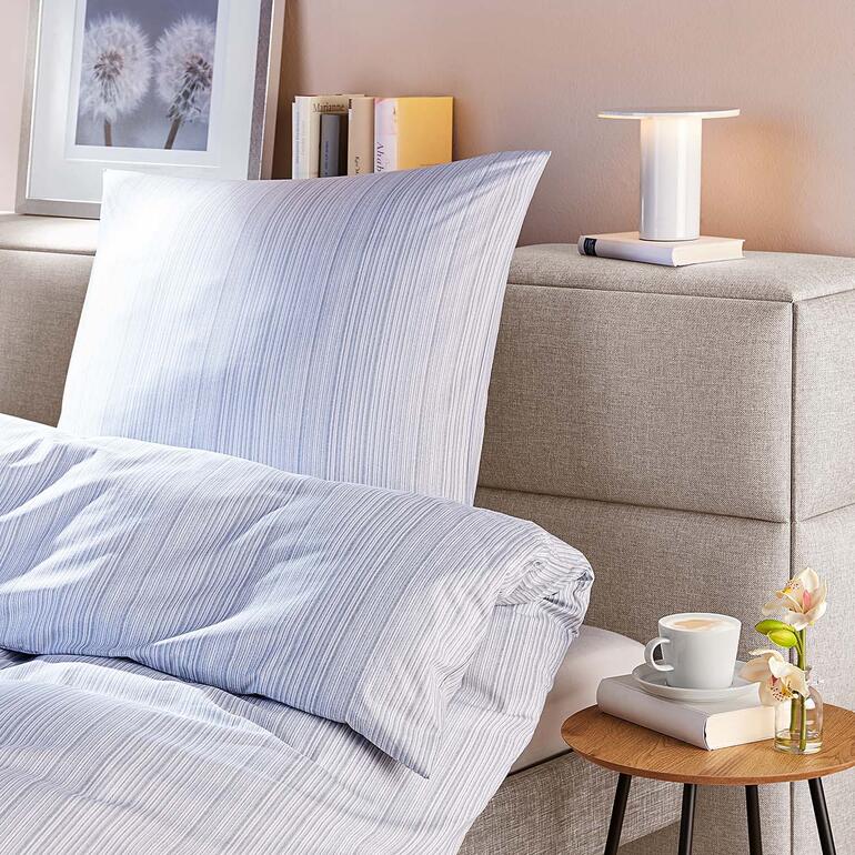 Bettwäsche mit Streifenmuster auf einem Bett mit grauem Kopfteil und moderner Leselampe.