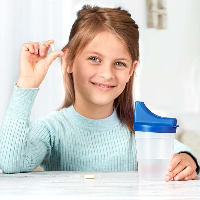Junge lächelt und hält eine Tablette in der Hand, vor einem Becher mit Wasser.