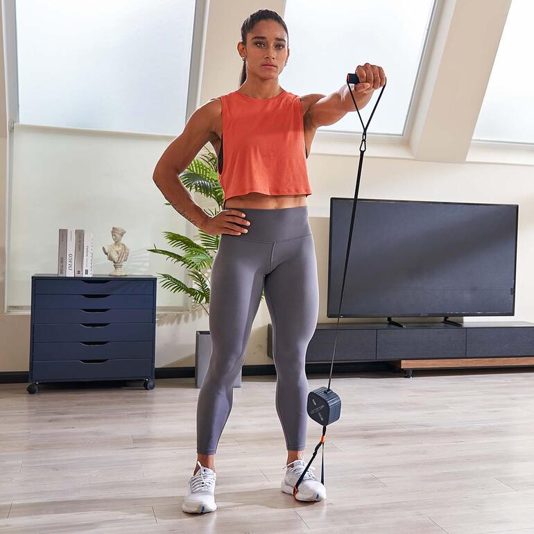 Frau mit blondem Haar und Sportoutfit, hält ein Pocket-Gym Gerät in einem modernen Raum.