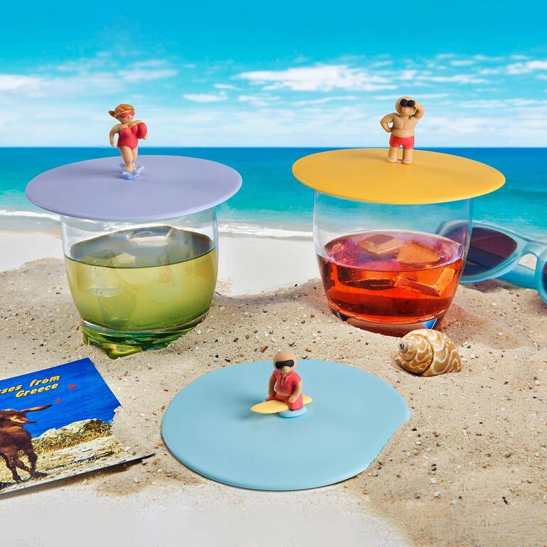 Drei Glasabdeckungen mit Surferfiguren stehen auf einem Sandstrand.