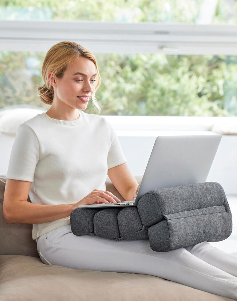 Frau sitzt mit Flexi-Rollenkissen, das ihre Arme unterstützt, am Laptop auf dem Sofa.