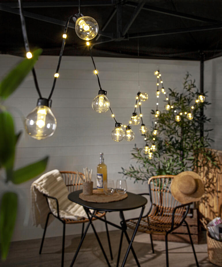 Beleuchtete Lichterkette über einem Tisch mit Stühlen im Außenbereich.