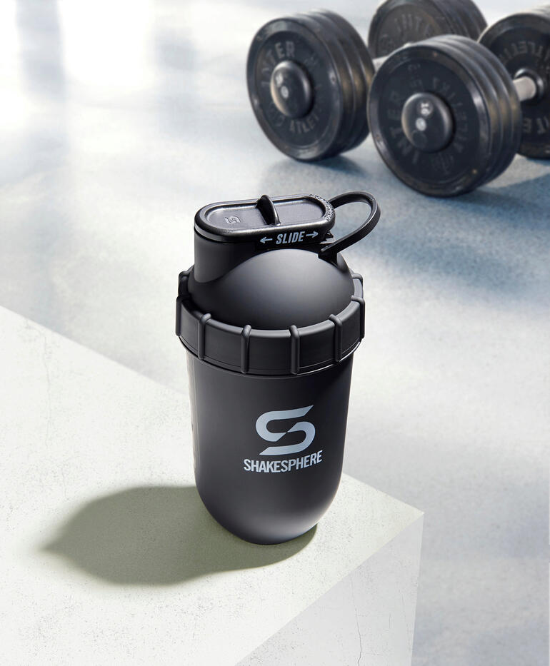 Dieser Fitness-Shaker nutzt allein die in der abgerundeten Kapselform entstehende Zentrifugalkraft – ohne Ecken, in denen sich Pulverrückstände festsetzen können. Schwarzer Shaker auf einer weißen Oberfläche mit Hanteln im Hintergrund.