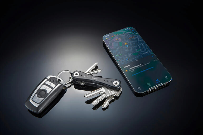 Wird Ihr Keymart® iPro geortet, sehen Sie die Position in der „Wo ist?“-App auf Ihrem iPhone. Smarter Schlüssel-Organizer neben einem Schlüsselbund und einem Smartphone auf einem Tisch.
