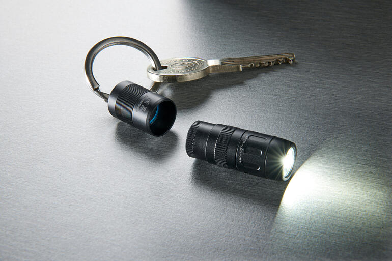 Einfach von der Schlüsselring-Kappe abziehen – und das Licht schaltet sich automatisch ein. Magnet-Mini-Taschenlampe und Schlüssel auf schimmernder Oberfläche, Lampe leuchtet.