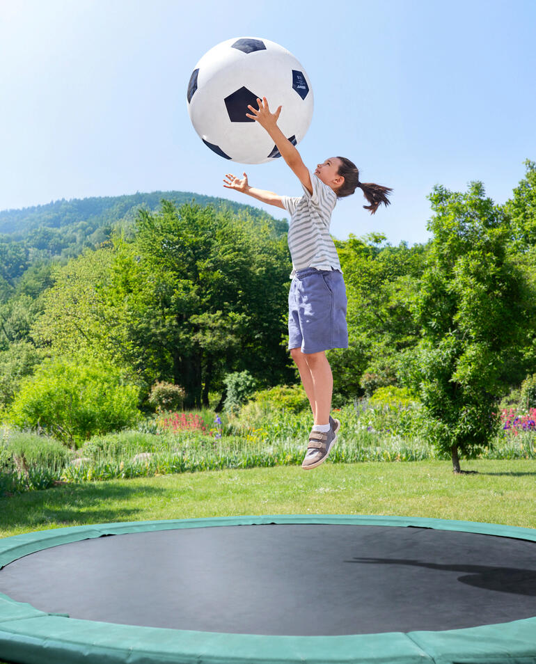 Riesiger Spielspaß auch auf dem Trampolin. Mädchen springt im Garten und greift nach einem großen Fußball.