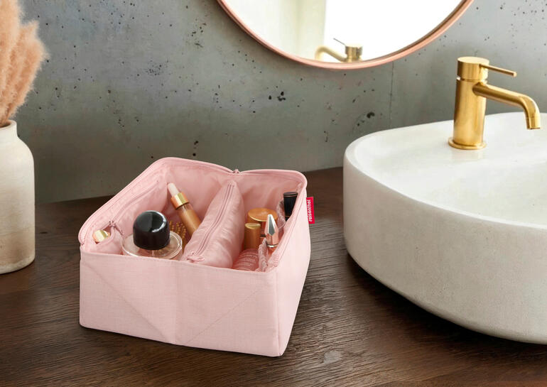 Viel Stauraum und praktische Aufteilung für beste Übersichtlichkeit. Offenes rosa 2-in-1 Reisenecessaire mit Pflegeprodukten auf einem Holztisch