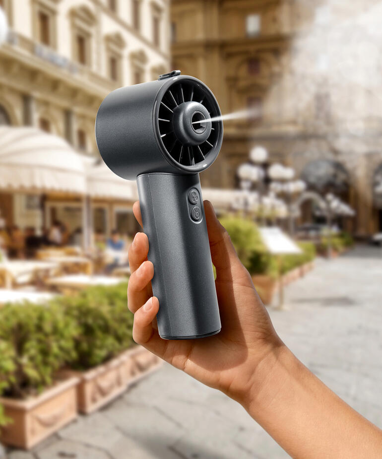Hand hält einen Hydro-Mini-Ventilator in einer Stadt, mit historischen Gebäuden im Hintergrund.