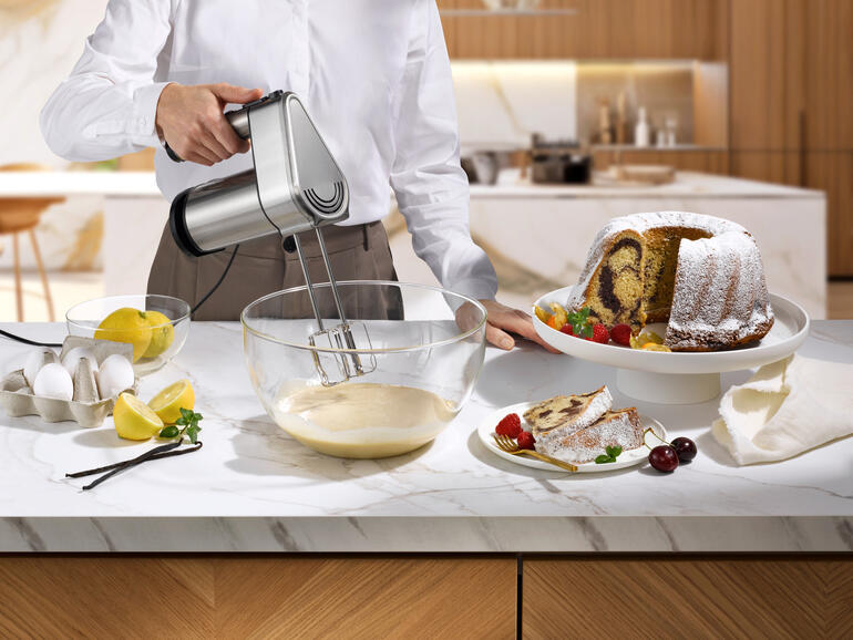 Mann hält Gastroback Power-Handmixer über einer Schüssel mit Teig und Kuchen auf einem Tisch.