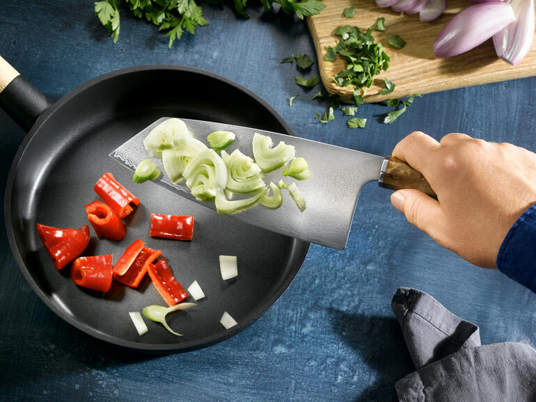 Hand mit Damast Nakiri-Messer, das Gemüse in einer Pfanne schneidet.