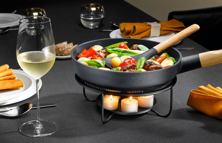 Pfanne mit Gemüse auf einem Stövchen, neben einem Weinglas auf einem gedeckten Tisch.