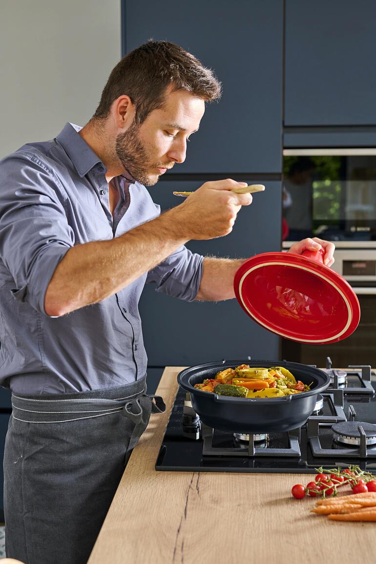 Mann in Küche, der Tajine Peugeot Saveurs mit einer Gabel probiert, während er den Deckel anhebt.