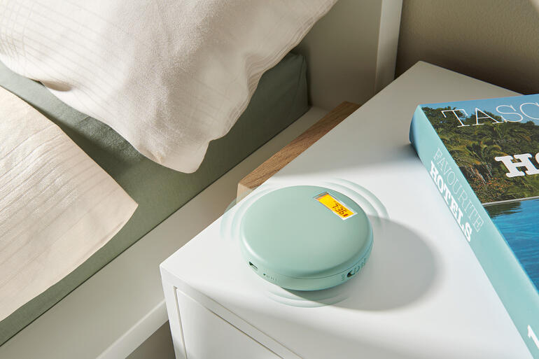 Vibrationswecker in sanftem Grün, auf einem Nachttisch neben einem Buch und einer Bettdecke.