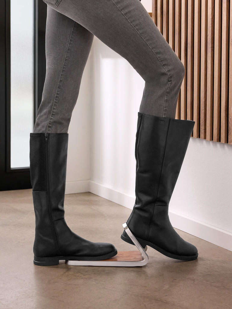 Frau benutzt einen Stiefelknecht, um ihre schwarzen Stiefel auszuziehen.