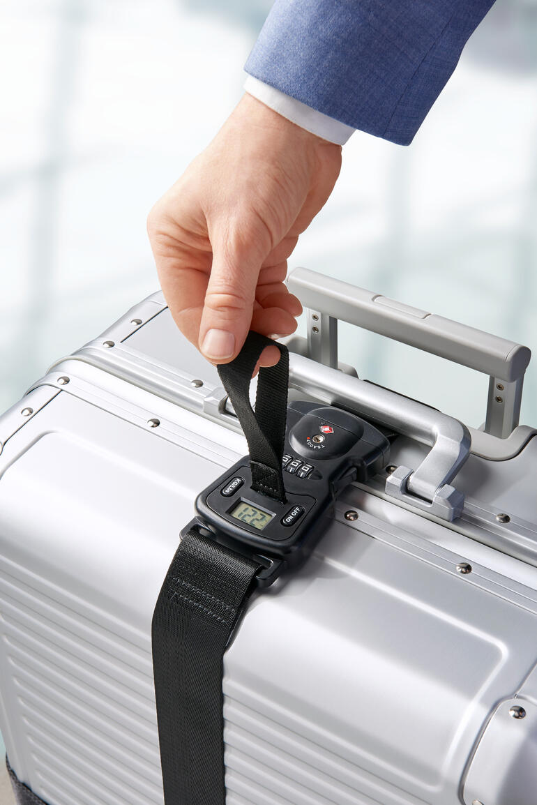 Person befestigt einen Koffergurt an einem silbernen Koffer am Flughafen.