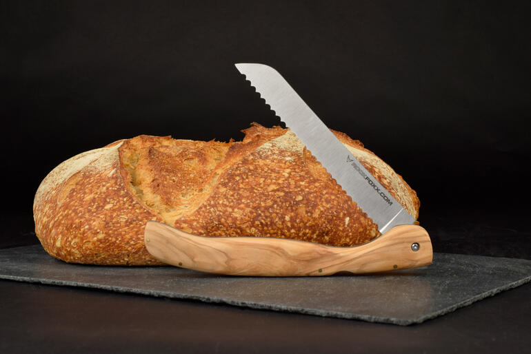 Klappbares Brotmesser mit Olivenholzgriff neben einem Laib Brot auf einer schwarzen Unterlage.