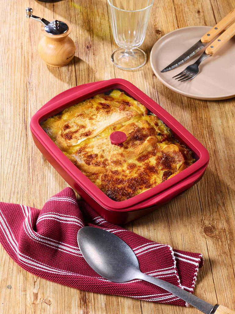 Rote Ofenform mit Deckel, gefüllt mit einer Bohnenspeise, auf einem Holztisch.