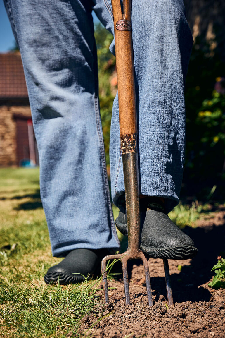 Person steht mit Gartenschaufel, trägt schwarze Airmesh Gartenschuhe.