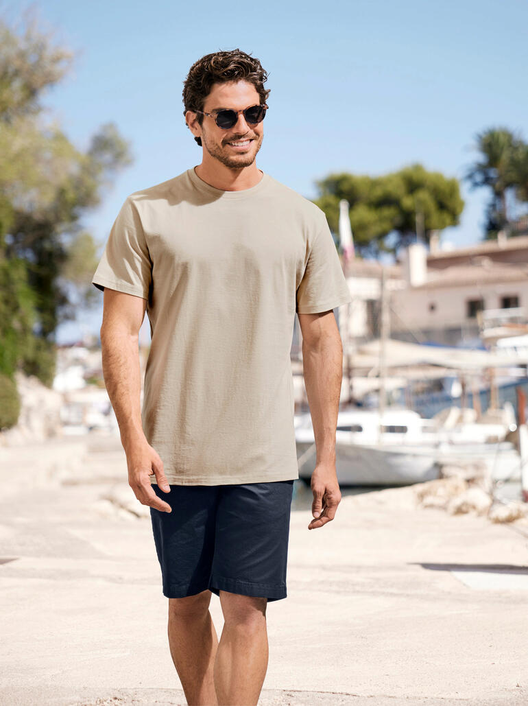 Lachender Mann im beigen T-Shirt und blauen Shorts bei einem Spaziergang am Hafen.