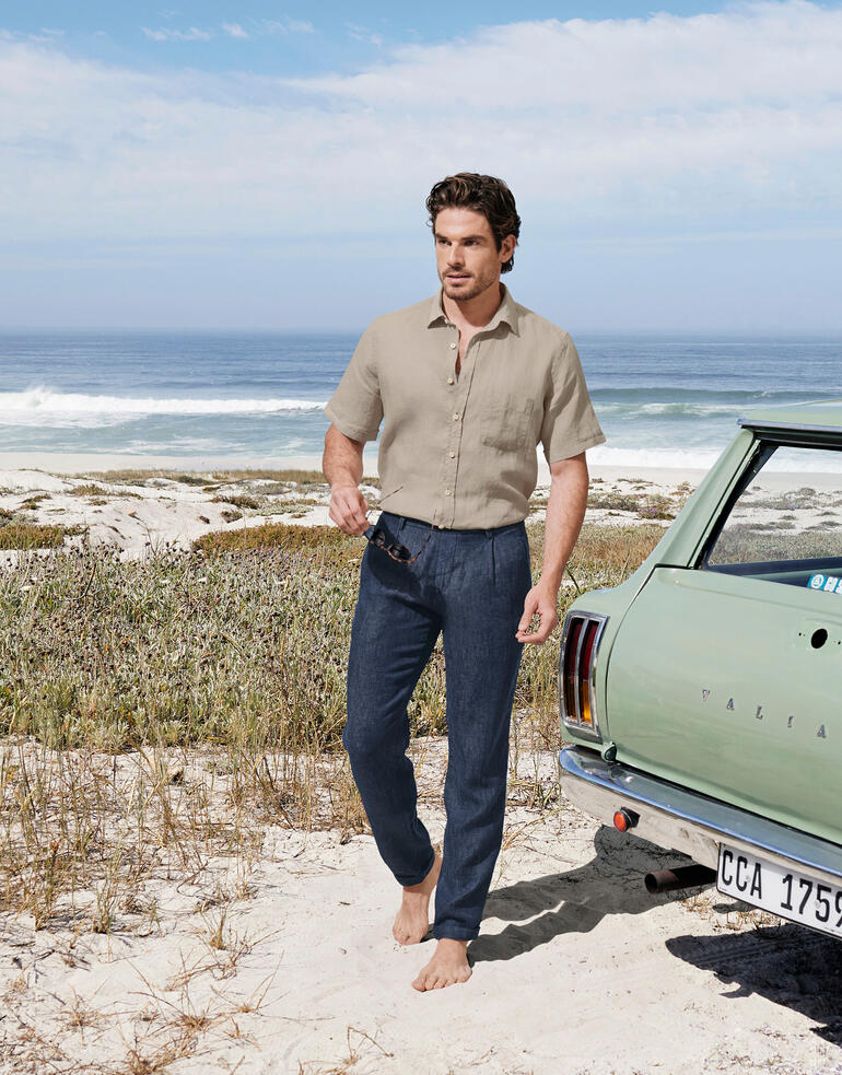 g1920 Indigo-Leinenhose Mann mit kahler Lederjacke und Indigo-Leinenhose am Strand vor einem Auto.